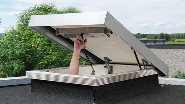 Man opening flat roof hatch on roof of house with a view of green trees and blue sky 